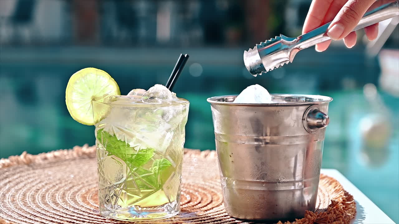 Cocktail and bucket with ice, slice of lime, man adding ice using tongs, pool on the background, Greece. Slow motion