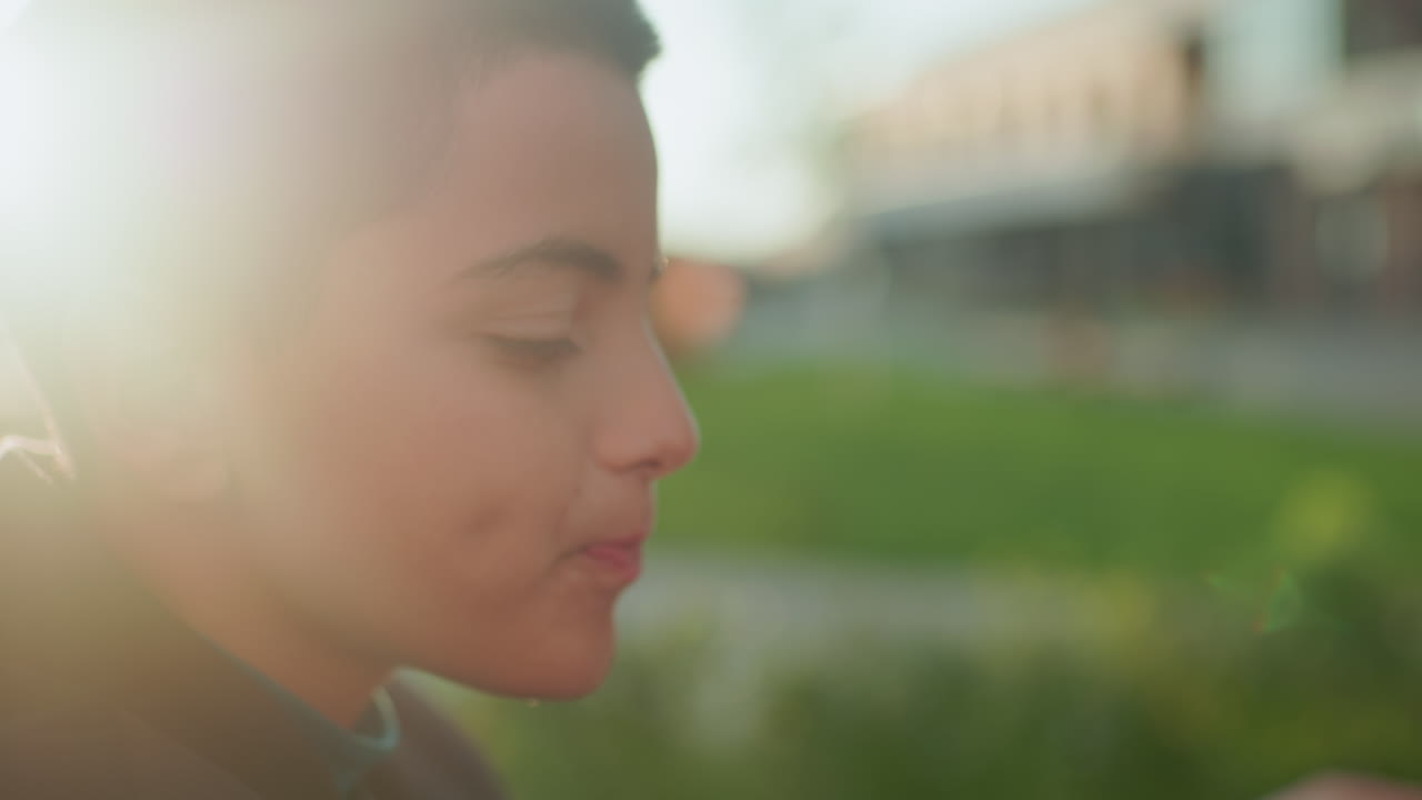 Close up side view of kid drinking grape juice from glass bottle outdoors, cheek dimples visible as he sips beverage under warm natural sunlight, enjoying refreshing drink in casual urban setting