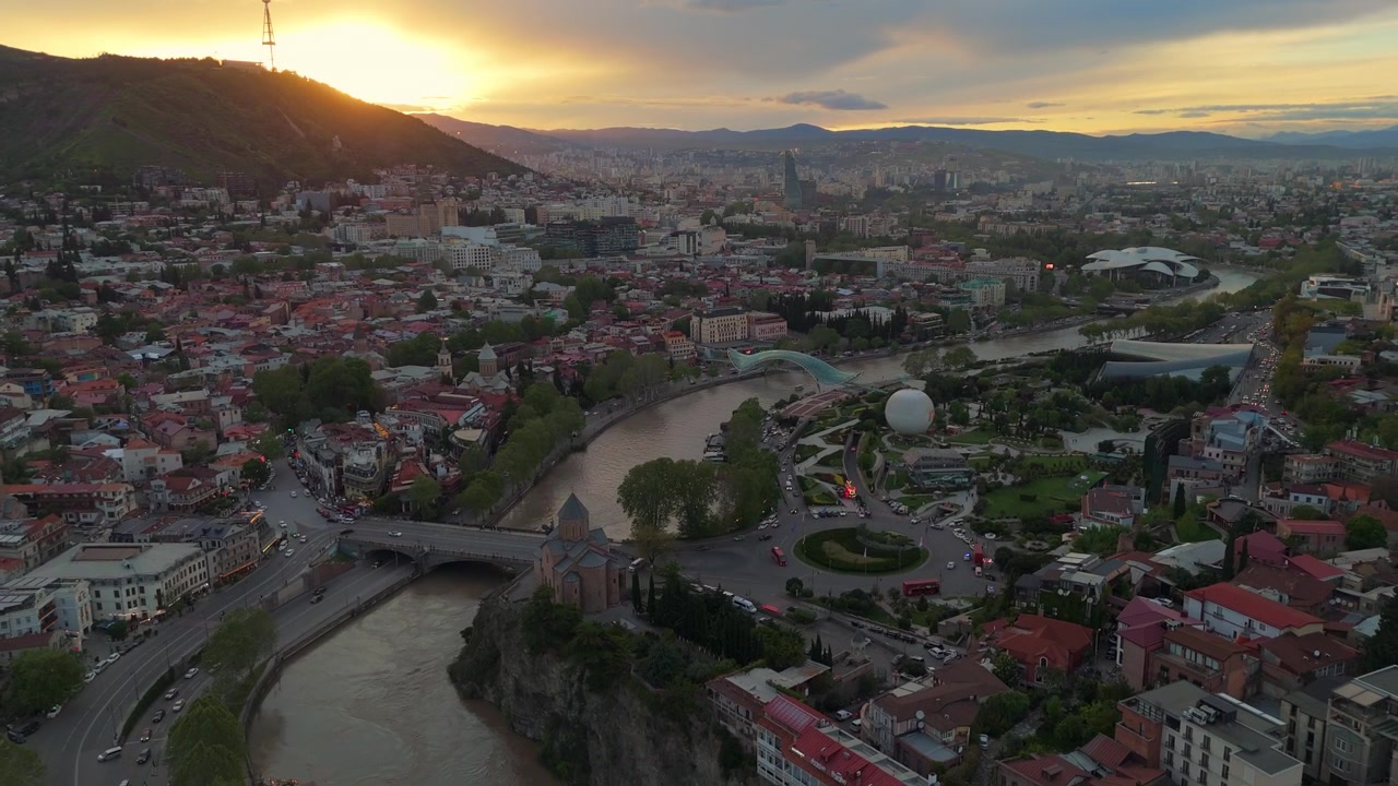 Letecký pohled na Tbilisi při západu slunce s Mostem míru, řekou Mtkvari a Halou veřejných služeb. Panorama hlavního města Gruzie s parkem Rike, kostelem Metekhi a historickým Starým Městem ve zlaté hodince.