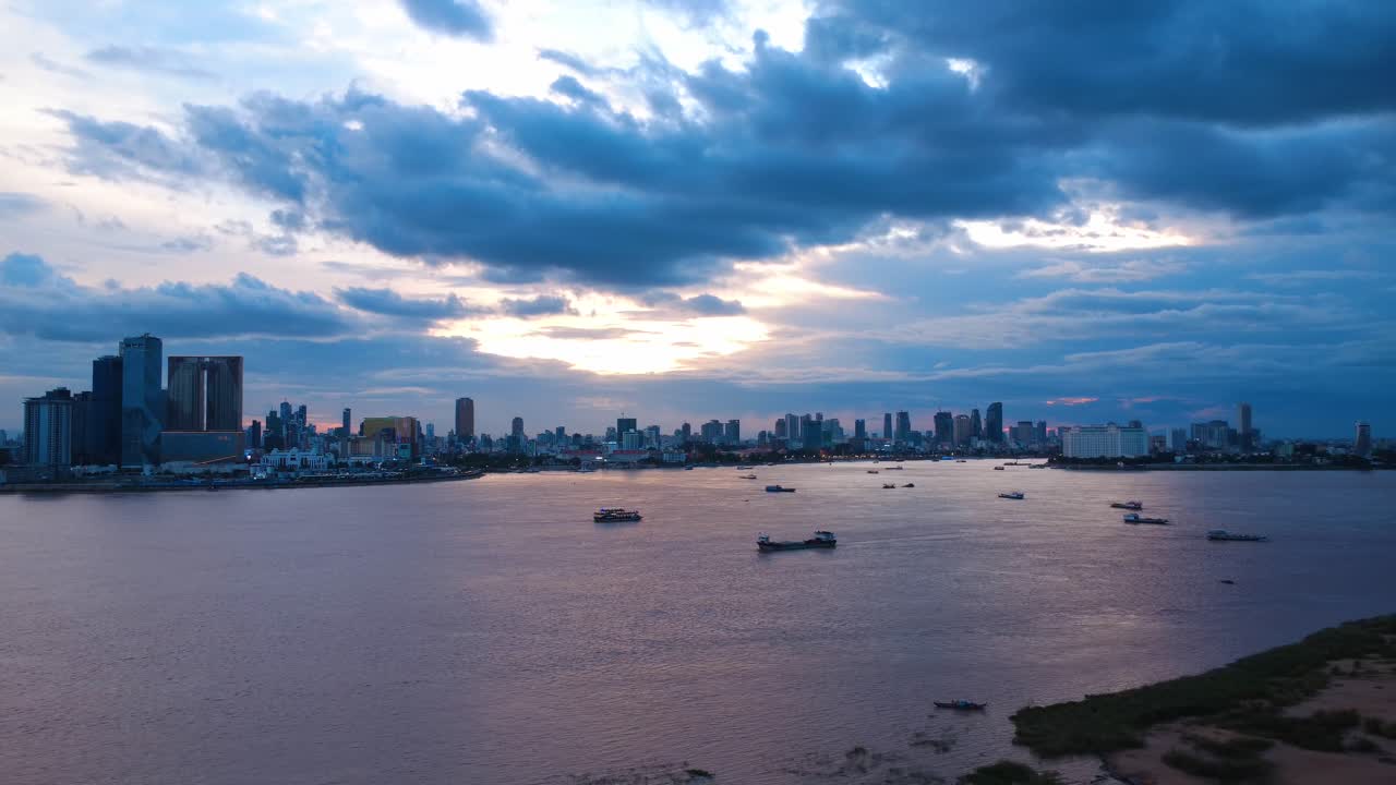 Breathtaking aerial footage of Phnom Penh's skyline at dusk, capturing the serene Mekong River under dramatic evening clouds. Vibrant cityscape with glowing lights reflects a tranquil,