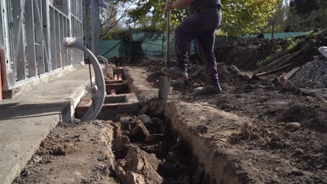 Close-up-shot-of-worker-ditching-with-a-shovel-around-a-steel-frame-house-at-construction-site-in-sunlight