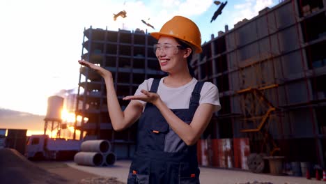 trabajadora asiática con gafas de protección y casco de seguridad sonriendo y señalando hacia un lado mientras está de pie en el sitio de construcción