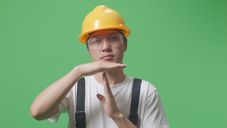 close up of asian man worker wearing goggles and safety helmet looking at camera and showing time out hands gesture while standing in the green screen background studio