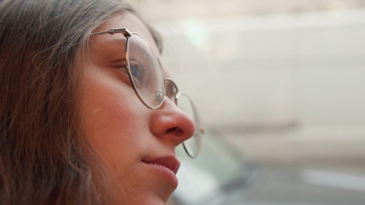 Donna riflessiva con occhiali osserva dall'interno dell'auto. Primo piano cinematografico di una donna con lentiggini all'interno di un veicolo. Signora introspettiva con occhiali rotondi che riflette silenziosamente all'interno di un'automobile in movimento.