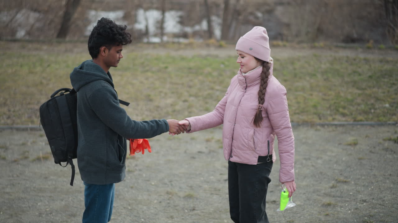 Black guy wearing dark gray hoodie holding orange gloves facing woman in pink jacket and beanie holding neon safety vest, standing outdoors in park during early spring with grassy background and cloudy