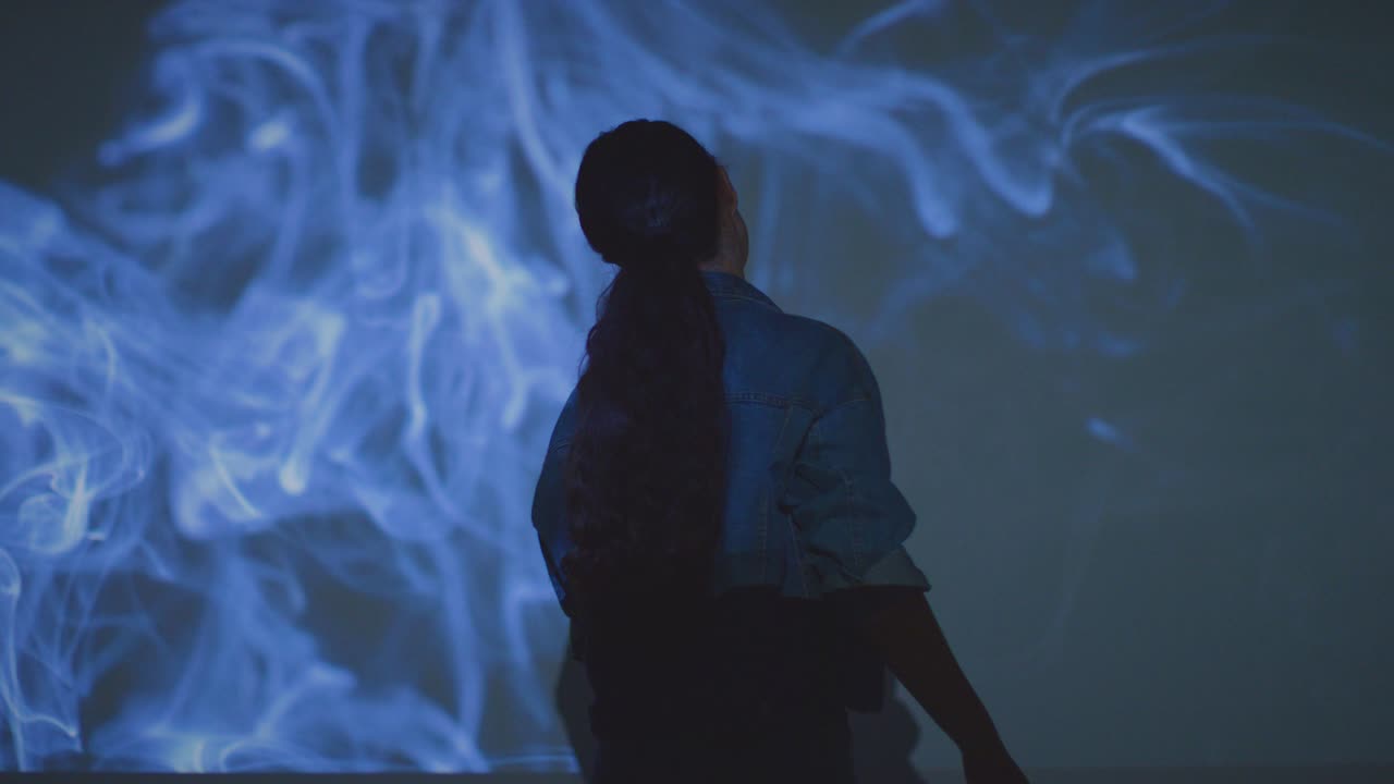 벽에 투사되는 연기의 이미지와 상호 작용하는 몰입 예술 전시회를 방문하는 여성