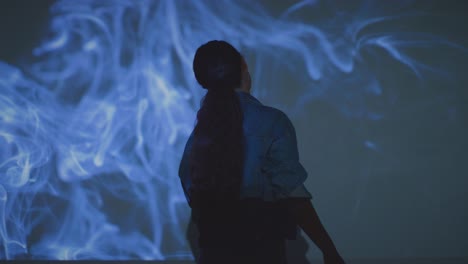 mujer visitando una exposición de arte inmersiva interactuando con imágenes de humo proyectadas en la pared