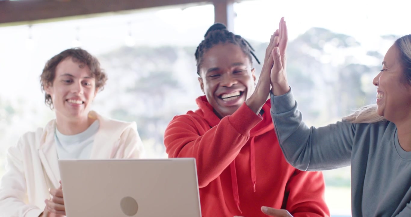 Diverse friends in hoodies leaning over silver laptop at cafe, pointing, clapping upon seeing win