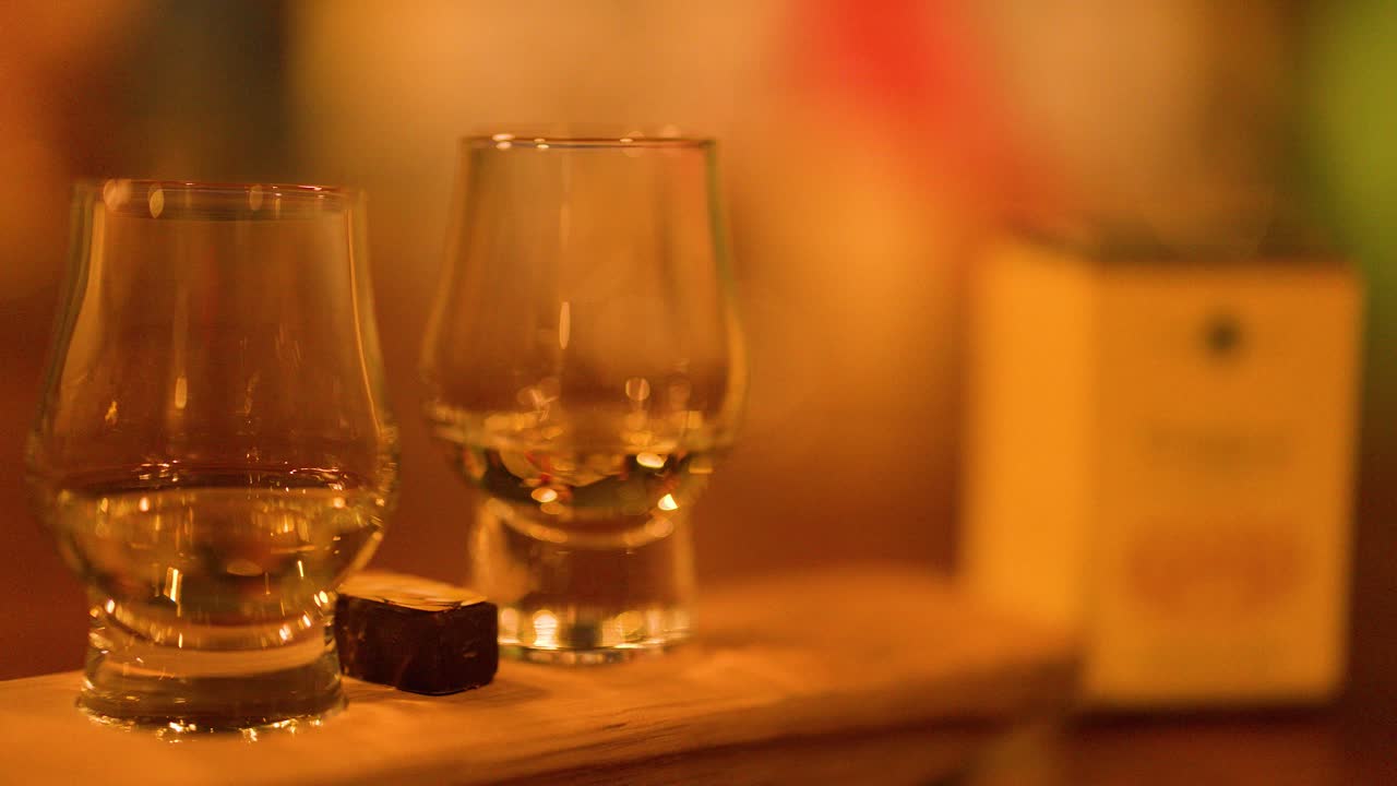 A hand lifts a whisky glass from a wooden tray in warm, ambient bar lighting