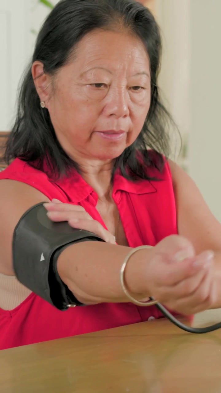 Video verticale: Anziano/a solleva il braccio sinistro e mette il bracciale a casa, collegando il tubo al monitor rosso per la pressione sanguigna.