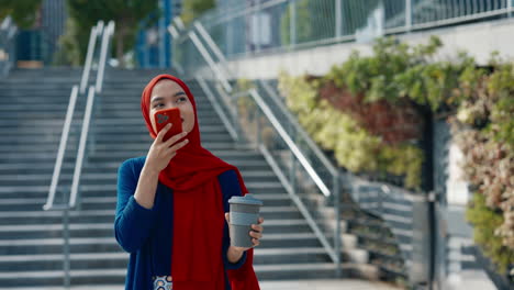 young muslim woman in a red hijab using a smartphone and holding a coffee cup outdoors