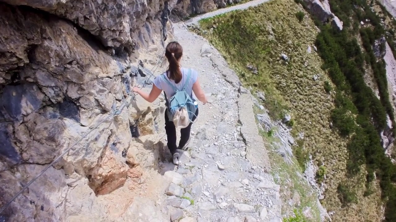 Giovane donna che fa escursioni su un sentiero stretto e protetto sul fianco di una montagna, aggrappata a un cavo di sicurezza con una profonda valle sottostante