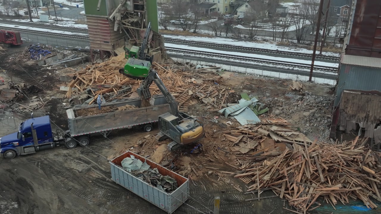 Trucks move to and from a demolition site of an old feed mill as machinery loads logs and wood debris. Workers carry out tasks in cold weather at the site. The ground is covered with piles of wood.