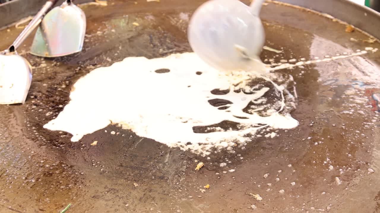 A ladle pours batter onto a hot griddle, spreading it evenly to form a crepe.