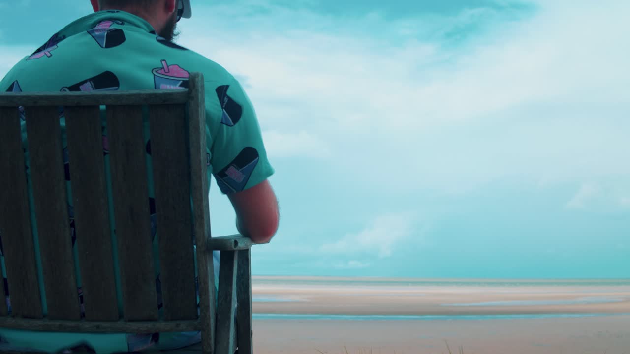 un joven se sienta en una sola silla de madera y observa la playa durante la marea baja bañada por la cálida luz del sol mientras una tormenta comienza a gestarse en el horizonte