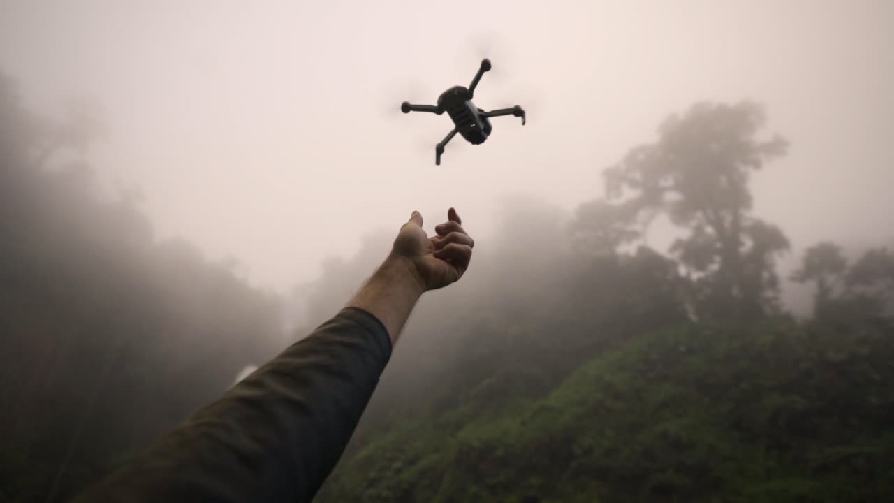 Drone descending toward a person's outstretched hand, against a backdrop of dense fog and the lush rainforest of Catarata del Toro