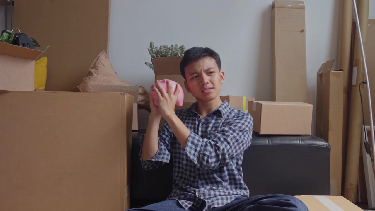Young man holds piggy bank while surrounded by packed cardboard boxes in home