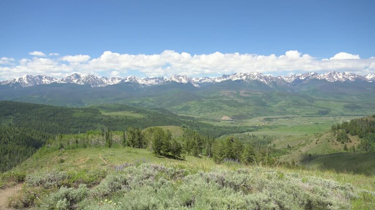 View from the top of a Colorado hiking trail overlooking a green valley with snowcapped mountains, static