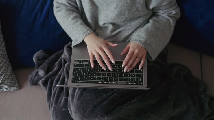 Nahaufnahme einer jungen Frau, die auf ihrem Laptop tippt, während sie bequem auf einem Sofa zu Hause sitzt. Die gemütliche Umgebung und die weiche Decke unterstreichen die Telearbeit und den modernen digitalen Lebensstil.