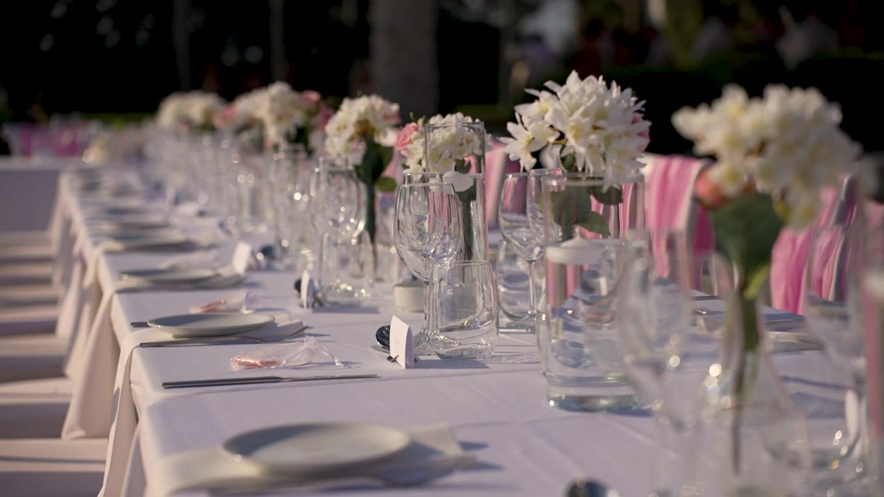 Et langt bord er dækket til et udendørs arrangement. Glas og tallerkener er pænt placeret, og blomster står i vaser. Opsætningen er klar til gæsterne i hyggelige omgivelser