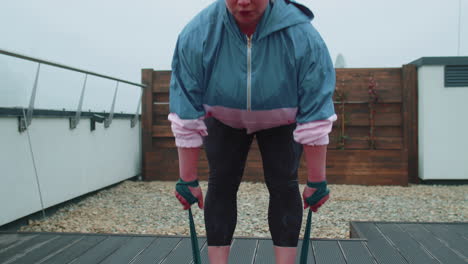 Sporty-girl-in-sportswear-does-yoga-stretching-exercising-using-rubber-band-on-roof-of-house-outdoor