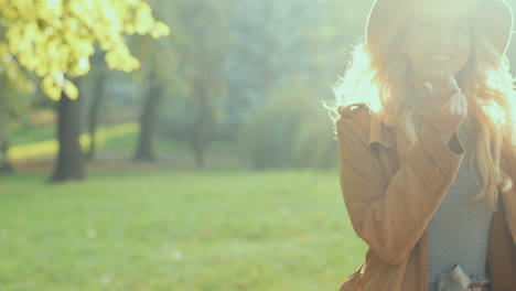 Vista-De-Cerca-De-Una-Joven-Rubia-Sosteniendo-Su-Sombrero-Y-Sonriendo-A-La-Cámara-En-El-Parque-En-Otoño