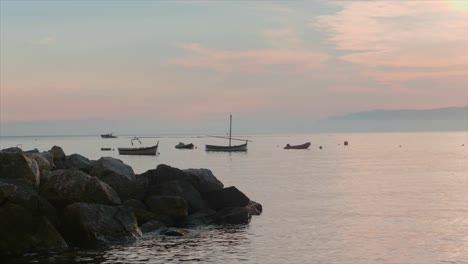 Still-boats-at-sunset,-a-speed-boat-moves-from-right-to-left,-rock-in-the-foreground,-shot-near-Portofinoo