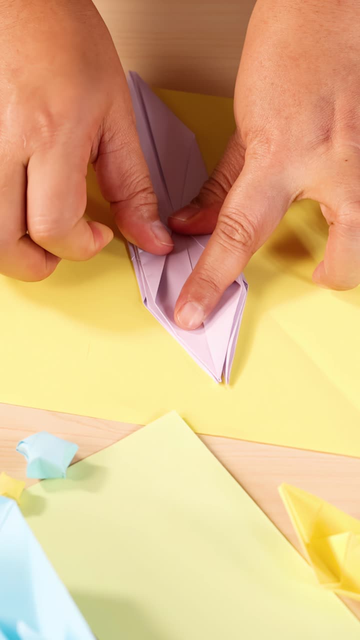Person carefully folds purple origami paper on yellow background, close-up, bright even lighting