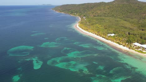 Vista-Aérea-De-La-Playa-Tropical-De-Arena-Blanca,-Arrecifes-De-Coral-Y-Agua-De-Mar-Turquesa-Clara-Con-Pequeñas-Olas-Y-Bosque-De-Palmeras