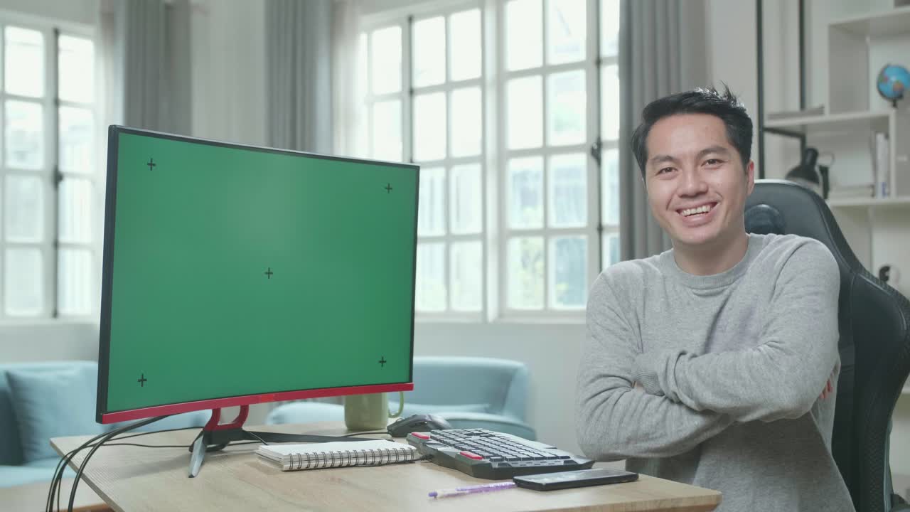 Asian Man In Long Sleeved T-Shirt With Green Screen Desktop Computer Crossing His Arms And Smiling To Camera While Working At Home.