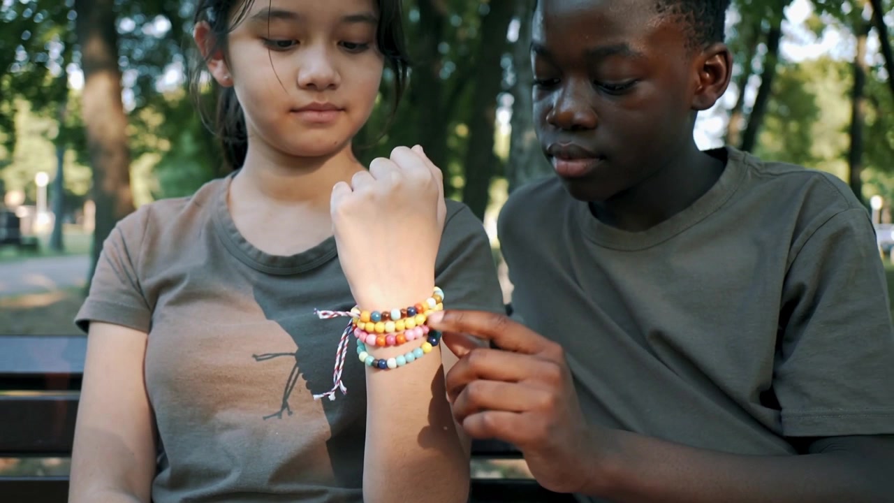 Hand entering frame, teens in olive shirts leaning in and checking colorful bracelets on park bench