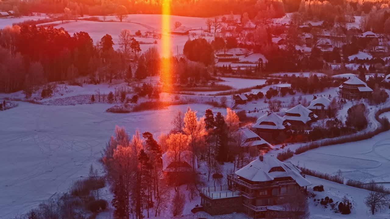 Dronekuva jäätyneen järven yllä talvisen auringonlaskun aikaan. Auringon häikäisy valaisee huurteisia puita ja lumista kylää. Lämmin oranssi taivas, rauhallinen maaseudun panoraama ja tilaa tekstille.