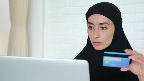 검은 히자브를 입은 여성이 노트북에 앉아 신용카드와 노트북을 사용하여 온라인으로 결제를 수행합니다. 무슬림 여성은 쇼핑과 집에서 상품을 주문하기 위해 인터넷을 사용합니다.