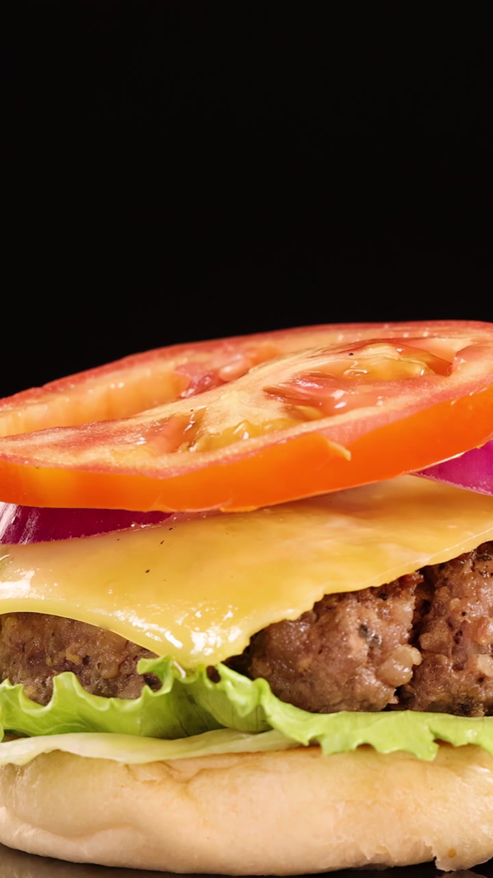 La parte superiore di un panino con semi di sesamo cade su un cheeseburger di manzo impilato con lattuga, pomodoro, cipolla e formaggio, ripreso al rallentatore su uno sfondo nero.