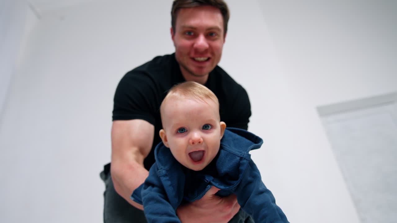 Lovely cute kid in hands of his dad. Father taking his baby boy close to the camera. Low angle view. White backdrop.