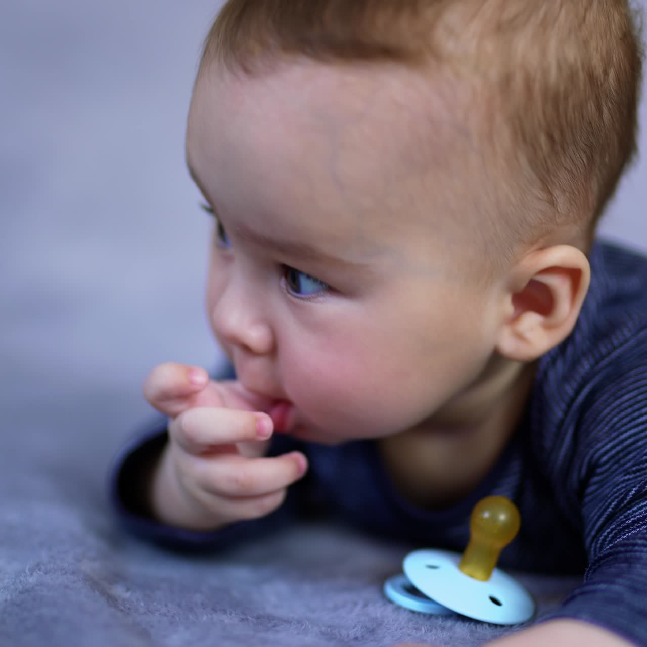 Sweet Caucasian toddler boy lying on belly with a pacifier in front of him. Cute kid looks aside holding his thumb in mouth