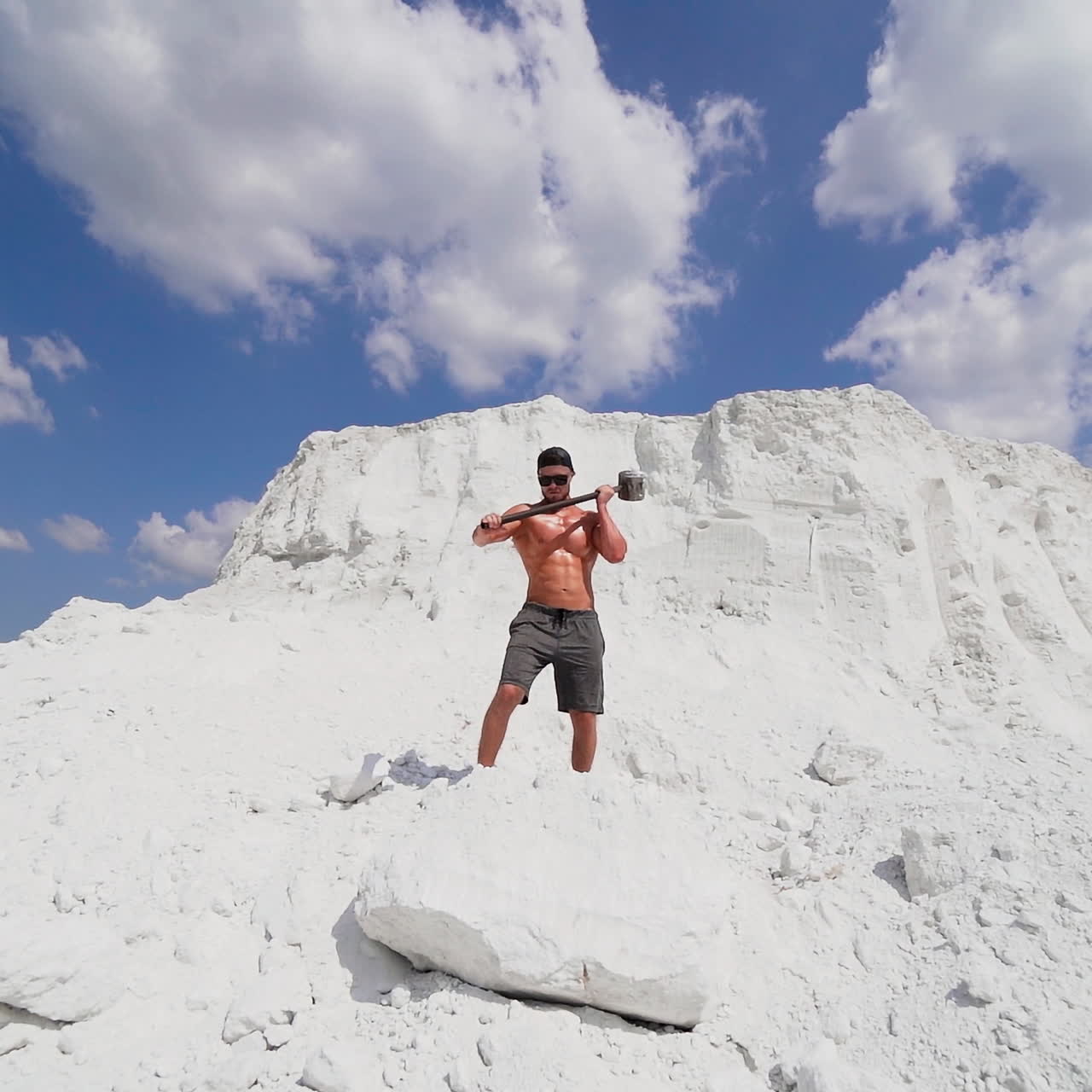 Muscular man on the background of white mountain. Shirtless guy with iron hammer on the natural background of large mountain in summer.