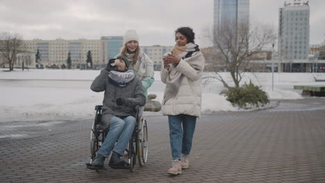 disabled man in wheelchair and two women walking together and drinking takeaway coffee in the city in winter