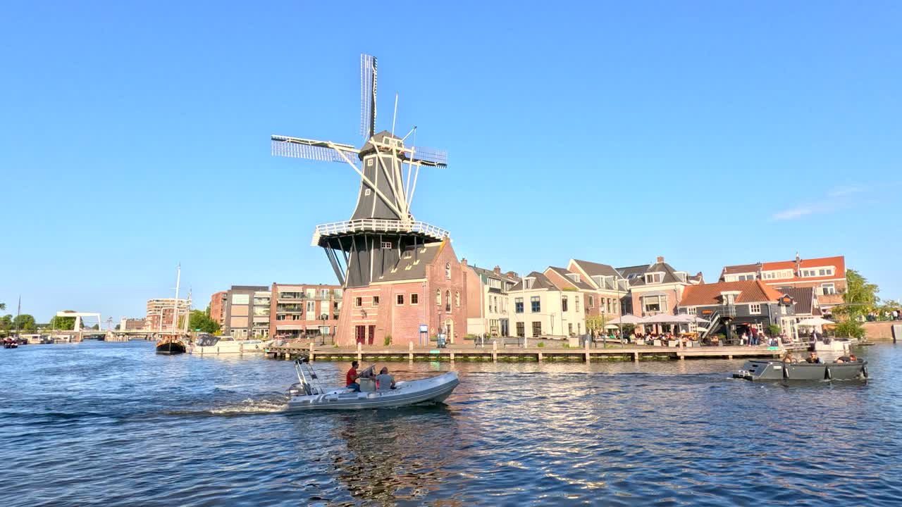 Motorboat travels across canal in Haarlem, passing windmill and historic buildings under clear daylight