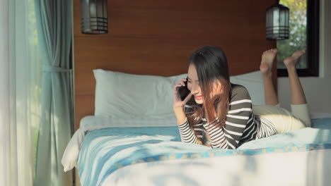 young asian woman talking on a mobile phone while lying on a bed on her stomach with her feet up