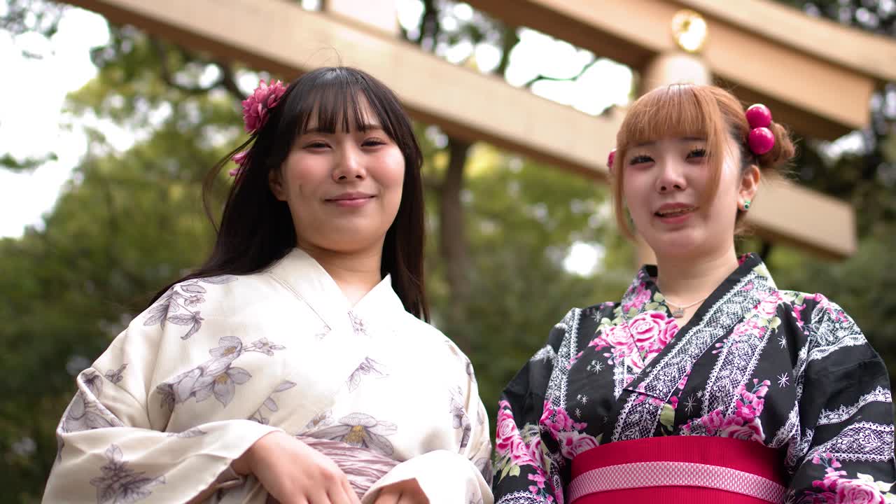 Toma en contrapicado de dos mujeres japonesas sonrientes vistiendo kimonos frente a una puerta torii de madera.
