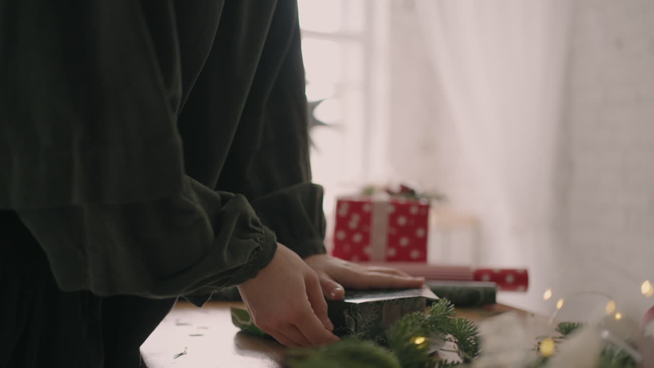 vista lateral de armar y preparar un regalo de navidad. un primer plano de una mujer envuelve sus manos alrededor de una caja en papel de regalo con un adorno navideño. envoltorio de regalo decorador profesional