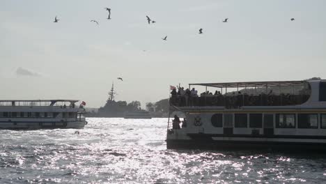 a ferry full of people sails on a river in istanbul with gulls flying in the air.