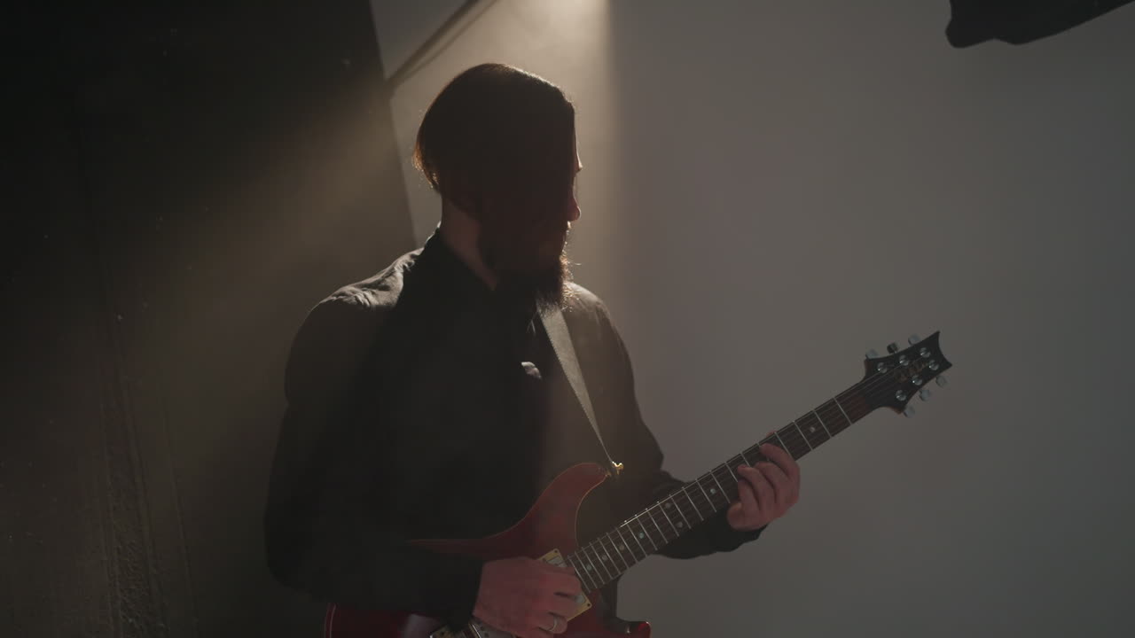 Bearded guitarist in black passionately playing a red guitar, head subtly moving in a dimly lit room