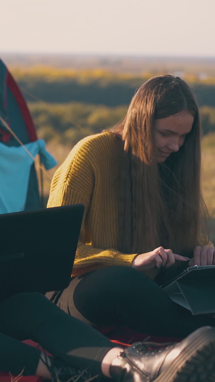beautiful girls use tablet and laptop working as freelancers sitting by blue tent on river bank in windy autumn evening