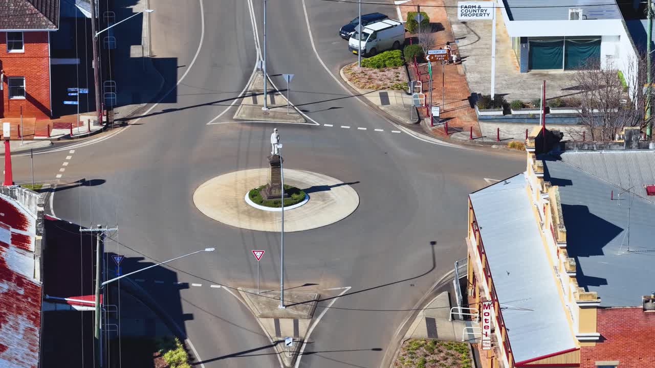 Drone ascends above quiet roundabout, revealing rural town streets, cars, shops, and bright daylight