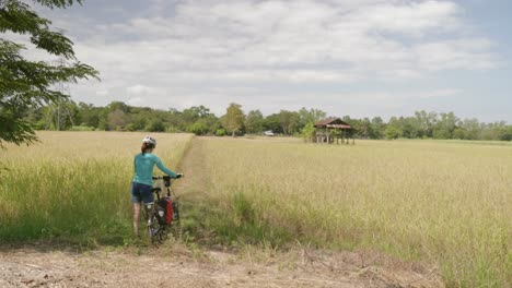 a stationary footage of a lone female cyclist walking her bike going into a rice farm or wheat field