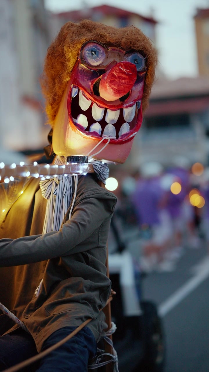 Testa di burattino spaventosa con naso rosso, grandi denti e occhi blu, parte di una sfilata di carnevale di sera.
