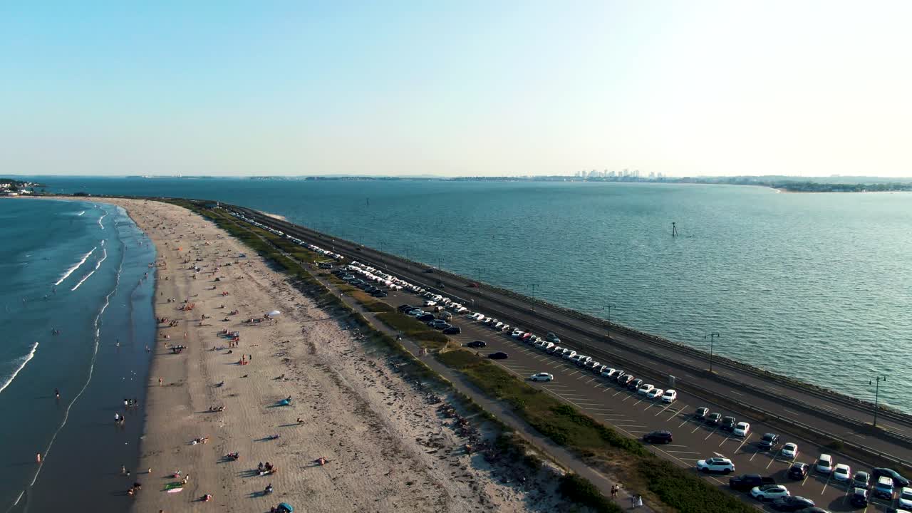 Nahant Beach full of people, aerial drone fly backward view. City skyline in far distance