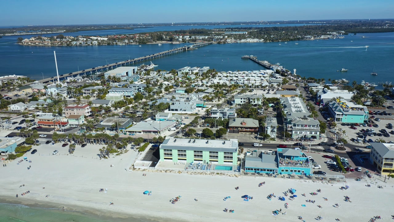 Aerial footage of a coastal town with a sandy beach and turquoise waters under a clear blue sky.